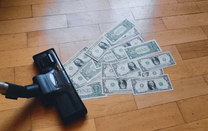 A vacuum sucking up money fanned out across a wood floor.